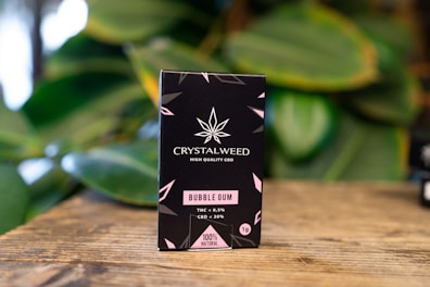 A black box of Crystalweed CBD product is placed on a wooden surface with a blurred background of green leaves. The packaging features a cannabis leaf design and text in pink and white, highlighting the product's flavor as bubble gum and its natural ingredients.