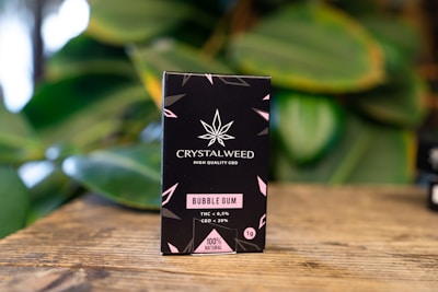 A black box of Crystalweed CBD product is placed on a wooden surface with a blurred background of green leaves. The packaging features a cannabis leaf design and text in pink and white, highlighting the product's flavor as bubble gum and its natural ingredients.