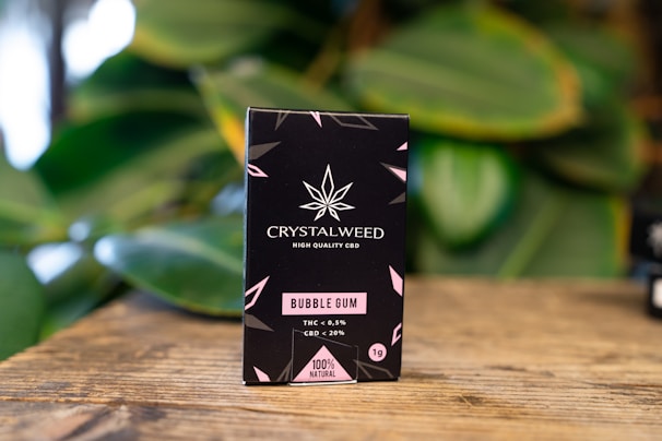 A black box of Crystalweed CBD product is placed on a wooden surface with a blurred background of green leaves. The packaging features a cannabis leaf design and text in pink and white, highlighting the product's flavor as bubble gum and its natural ingredients.