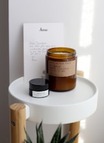 A handwritten note and two products are arranged on a small, modern round tray with wooden legs. The note features elegant lettering at the top. There is a brown glass jar labeled as a soy candle and a smaller black and white container labeled as a lip balm.