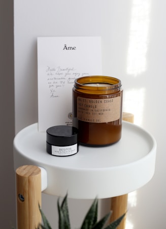 A handwritten note and two products are arranged on a small, modern round tray with wooden legs. The note features elegant lettering at the top. There is a brown glass jar labeled as a soy candle and a smaller black and white container labeled as a lip balm.