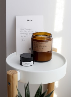 A handwritten note and two products are arranged on a small, modern round tray with wooden legs. The note features elegant lettering at the top. There is a brown glass jar labeled as a soy candle and a smaller black and white container labeled as a lip balm.