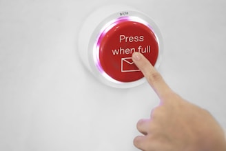 A hand is pressing a red button that has the text 'Press when full' and an envelope icon on it. The button is mounted on a white surface.