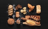 An assortment of various types of bread and pastries arranged on a dark surface. The selection includes a braided loaf sprinkled with seeds, a couple of flaky croissants, a rustic round bread, and several long baguettes. There are also smaller baked goods such as muffins and slices of dark, nutty bread. Some flour is scattered around, adding to the artisanal look.