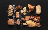 An assortment of various types of bread and pastries arranged on a dark surface. The selection includes a braided loaf sprinkled with seeds, a couple of flaky croissants, a rustic round bread, and several long baguettes. There are also smaller baked goods such as muffins and slices of dark, nutty bread. Some flour is scattered around, adding to the artisanal look.