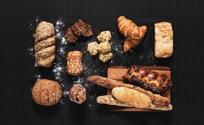 An assortment of various types of bread and pastries arranged on a dark surface. The selection includes a braided loaf sprinkled with seeds, a couple of flaky croissants, a rustic round bread, and several long baguettes. There are also smaller baked goods such as muffins and slices of dark, nutty bread. Some flour is scattered around, adding to the artisanal look.