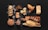 An assortment of various types of bread and pastries arranged on a dark surface. The selection includes a braided loaf sprinkled with seeds, a couple of flaky croissants, a rustic round bread, and several long baguettes. There are also smaller baked goods such as muffins and slices of dark, nutty bread. Some flour is scattered around, adding to the artisanal look.