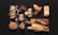 An assortment of various types of bread and pastries arranged on a dark surface. The selection includes a braided loaf sprinkled with seeds, a couple of flaky croissants, a rustic round bread, and several long baguettes. There are also smaller baked goods such as muffins and slices of dark, nutty bread. Some flour is scattered around, adding to the artisanal look.