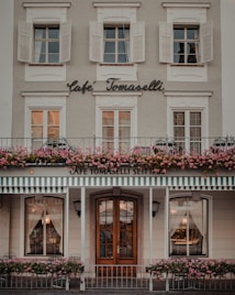 A charming cafe facade featuring a traditional design with white window shutters and a striped awning. Pink flowers line the balcony, adding a vibrant touch to the exterior. Wooden doors and elegant curtains are visible, contributing to a cozy and inviting atmosphere.