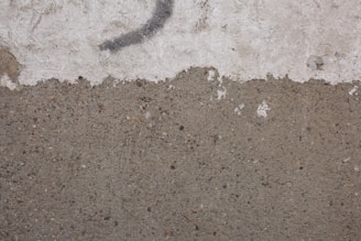 A textured wall with a rough, concrete surface on the lower half and a worn, white plaster section above. A faint, dark curved line runs across the upper portion, suggesting possible graffiti or a mark.