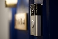 A smart video doorbell is mounted on a deep blue door with a blurred out number plate in the background.
