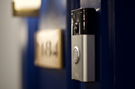 A smart video doorbell is mounted on a deep blue door with a blurred out number plate in the background.