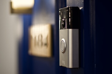 A smart video doorbell is mounted on a deep blue door with a blurred out number plate in the background.
