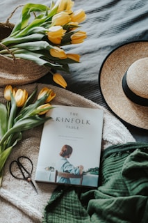A cozy setting featuring a straw hat, a bunch of yellow tulips in a woven basket, a thick green blanket fabric, a pair of scissors, and a book titled 'The Kinfolk Table' on a soft, textured backdrop.