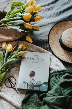 A cozy setting featuring a straw hat, a bunch of yellow tulips in a woven basket, a thick green blanket fabric, a pair of scissors, and a book titled 'The Kinfolk Table' on a soft, textured backdrop.