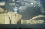 A car's windshield with a large circular crack and multiple radiating fractures. Inside the vehicle, beige leather seats are visible, along with a steering wheel. A blue air freshener shaped like a tree hangs from the rearview mirror.
