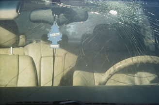 A car's windshield with a large circular crack and multiple radiating fractures. Inside the vehicle, beige leather seats are visible, along with a steering wheel. A blue air freshener shaped like a tree hangs from the rearview mirror.