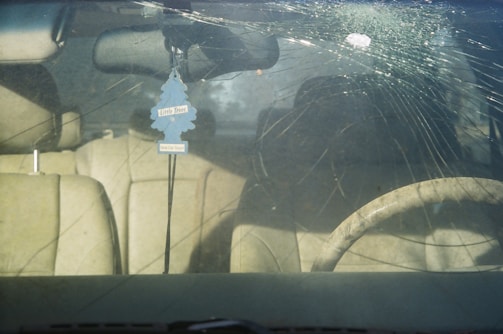A car's windshield with a large circular crack and multiple radiating fractures. Inside the vehicle, beige leather seats are visible, along with a steering wheel. A blue air freshener shaped like a tree hangs from the rearview mirror.