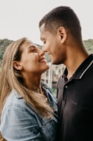 A couple stands facing each other closely, smiling warmly and looking into each other's eyes. They appear to be in an outdoor setting with greenery in the background. The woman has long blonde hair and is wearing a denim jacket, while the man has short dark hair and is wearing a black polo shirt.