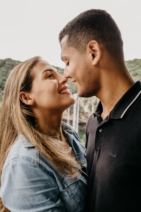 A couple stands facing each other closely, smiling warmly and looking into each other's eyes. They appear to be in an outdoor setting with greenery in the background. The woman has long blonde hair and is wearing a denim jacket, while the man has short dark hair and is wearing a black polo shirt.