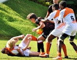 A group of young rugby players are engaged in a competitive match. The players are wearing different colored uniforms, with some in orange and white and others in black. One player is holding the rugby ball while being tackled by opponents. The scene takes place on a grassy field, and the action conveys a sense of intensity and teamwork.