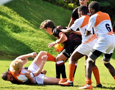 A group of young rugby players are engaged in a competitive match. The players are wearing different colored uniforms, with some in orange and white and others in black. One player is holding the rugby ball while being tackled by opponents. The scene takes place on a grassy field, and the action conveys a sense of intensity and teamwork.