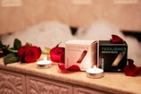 On a warmly lit, tiled surface, two boxes of Resilience CBD bath bombs are displayed, surrounded by red roses and small lit candles. The boxes are labeled 'Revive' and 'Recover', with petals scattered around them and bubbles faintly visible in the background.