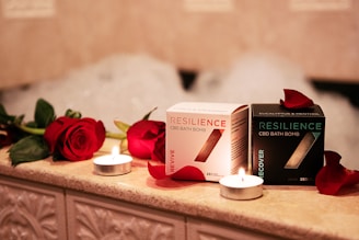 On a warmly lit, tiled surface, two boxes of Resilience CBD bath bombs are displayed, surrounded by red roses and small lit candles. The boxes are labeled 'Revive' and 'Recover', with petals scattered around them and bubbles faintly visible in the background.