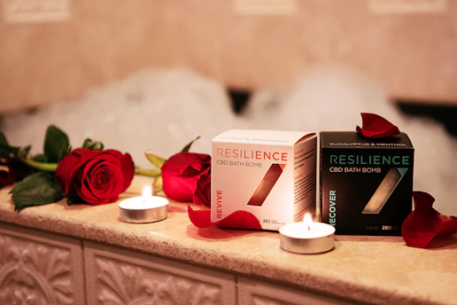 On a warmly lit, tiled surface, two boxes of Resilience CBD bath bombs are displayed, surrounded by red roses and small lit candles. The boxes are labeled 'Revive' and 'Recover', with petals scattered around them and bubbles faintly visible in the background.