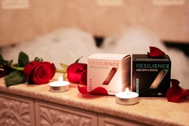 On a warmly lit, tiled surface, two boxes of Resilience CBD bath bombs are displayed, surrounded by red roses and small lit candles. The boxes are labeled 'Revive' and 'Recover', with petals scattered around them and bubbles faintly visible in the background.
