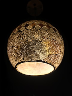 A spherical lamp with a mosaic-like texture made of small, irregular pieces, featuring earthy tones. The lamp emits a warm, soft glow against a dark background, creating a cozy and intimate ambiance.