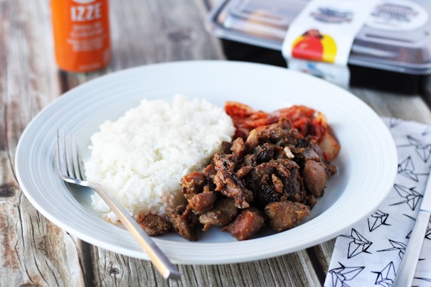 A plate of white rice, seasoned meat, and kimchi is arranged on a wooden table. Next to the plate is a fork, and beside it, there appears to be a patterned napkin. In the background, part of a carton and a bottle of orange Izze drink are visible.