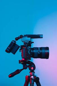 A professional camera mounted on a tripod against a gradient blue and pink background, with lighting creating a dramatic effect on the equipment.