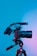 A professional camera mounted on a tripod against a gradient blue and pink background, with lighting creating a dramatic effect on the equipment.