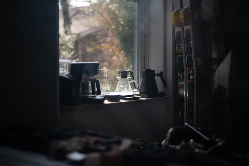A dimly lit kitchen scene with a window. Various coffee-related items, including a coffee pot, jar, and kettle, are placed on the windowsill. Light filters through the window, casting soft shadows across the surfaces. The background appears slightly blurred, suggesting a cozy and serene atmosphere.