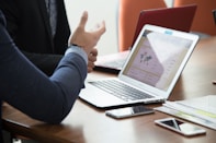 Two people are sitting at a wooden table, engaging in a discussion. A laptop is open, displaying a statistical dashboard with charts and a world map. Several smartphones and notepads are scattered on the table. The focus is on the person's hand gesturing in conversation, indicating active engagement.