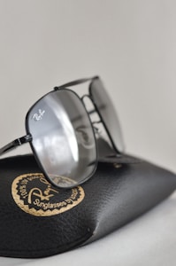 A pair of glasses resting on a textured black case with a distinctive circular golden logo. The lenses have a reflective surface, and the frame is sleek and stylish.