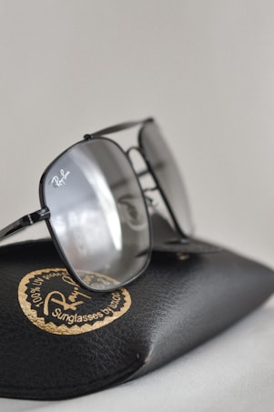 A pair of glasses resting on a textured black case with a distinctive circular golden logo. The lenses have a reflective surface, and the frame is sleek and stylish.