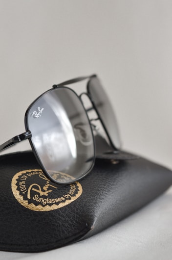 A pair of glasses resting on a textured black case with a distinctive circular golden logo. The lenses have a reflective surface, and the frame is sleek and stylish.