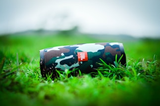 A camouflaged portable speaker rests on vibrant green grass, with a blurred natural background. The speaker's design features a mix of green, brown, and black hues, enhancing its outdoor appearance.