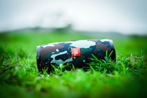 A camouflaged portable speaker rests on vibrant green grass, with a blurred natural background. The speaker's design features a mix of green, brown, and black hues, enhancing its outdoor appearance.