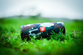 A camouflaged portable speaker rests on vibrant green grass, with a blurred natural background. The speaker's design features a mix of green, brown, and black hues, enhancing its outdoor appearance.
