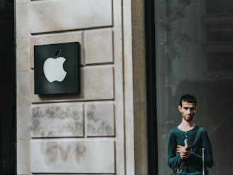 A man stands outside a building with a prominent Apple logo on the wall next to him. The building facade is made of light stone with a modern design. The man is casually dressed, carrying a bag with a shoulder strap, and looking at a mobile device. The area is well-lit, suggesting it might be daytime.
