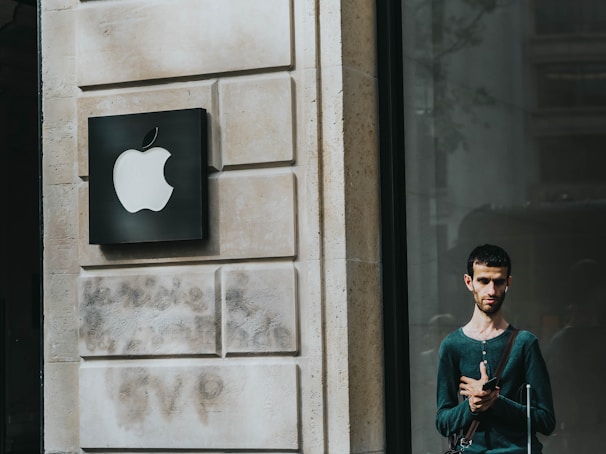 A man stands outside a building with a prominent Apple logo on the wall next to him. The building facade is made of light stone with a modern design. The man is casually dressed, carrying a bag with a shoulder strap, and looking at a mobile device. The area is well-lit, suggesting it might be daytime.