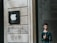 A man stands outside a building with a prominent Apple logo on the wall next to him. The building facade is made of light stone with a modern design. The man is casually dressed, carrying a bag with a shoulder strap, and looking at a mobile device. The area is well-lit, suggesting it might be daytime.