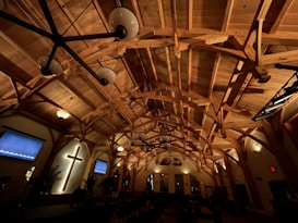 A wooden church interior featuring exposed beams and a prominent cross on the wall. The lighting is warm and dim, casting subtle shadows across the wooden structure. Several pendant lights hang from the ceiling, and there are screens on either side of the altar area. Plants and decorations are sparsely placed around the room.