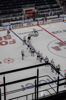A hockey team is lined up on the ice rink with players wearing white jerseys and dark pants. Some players are positioned in a skating stance, holding hockey sticks. A goalie in red gear is near the net, and the stands in the background appear empty. Advertisements and signage are visible on the rink boards.