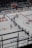 A hockey team is lined up on the ice rink with players wearing white jerseys and dark pants. Some players are positioned in a skating stance, holding hockey sticks. A goalie in red gear is near the net, and the stands in the background appear empty. Advertisements and signage are visible on the rink boards.