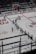 A hockey team is lined up on the ice rink with players wearing white jerseys and dark pants. Some players are positioned in a skating stance, holding hockey sticks. A goalie in red gear is near the net, and the stands in the background appear empty. Advertisements and signage are visible on the rink boards.