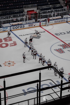 A hockey team is lined up on the ice rink with players wearing white jerseys and dark pants. Some players are positioned in a skating stance, holding hockey sticks. A goalie in red gear is near the net, and the stands in the background appear empty. Advertisements and signage are visible on the rink boards.